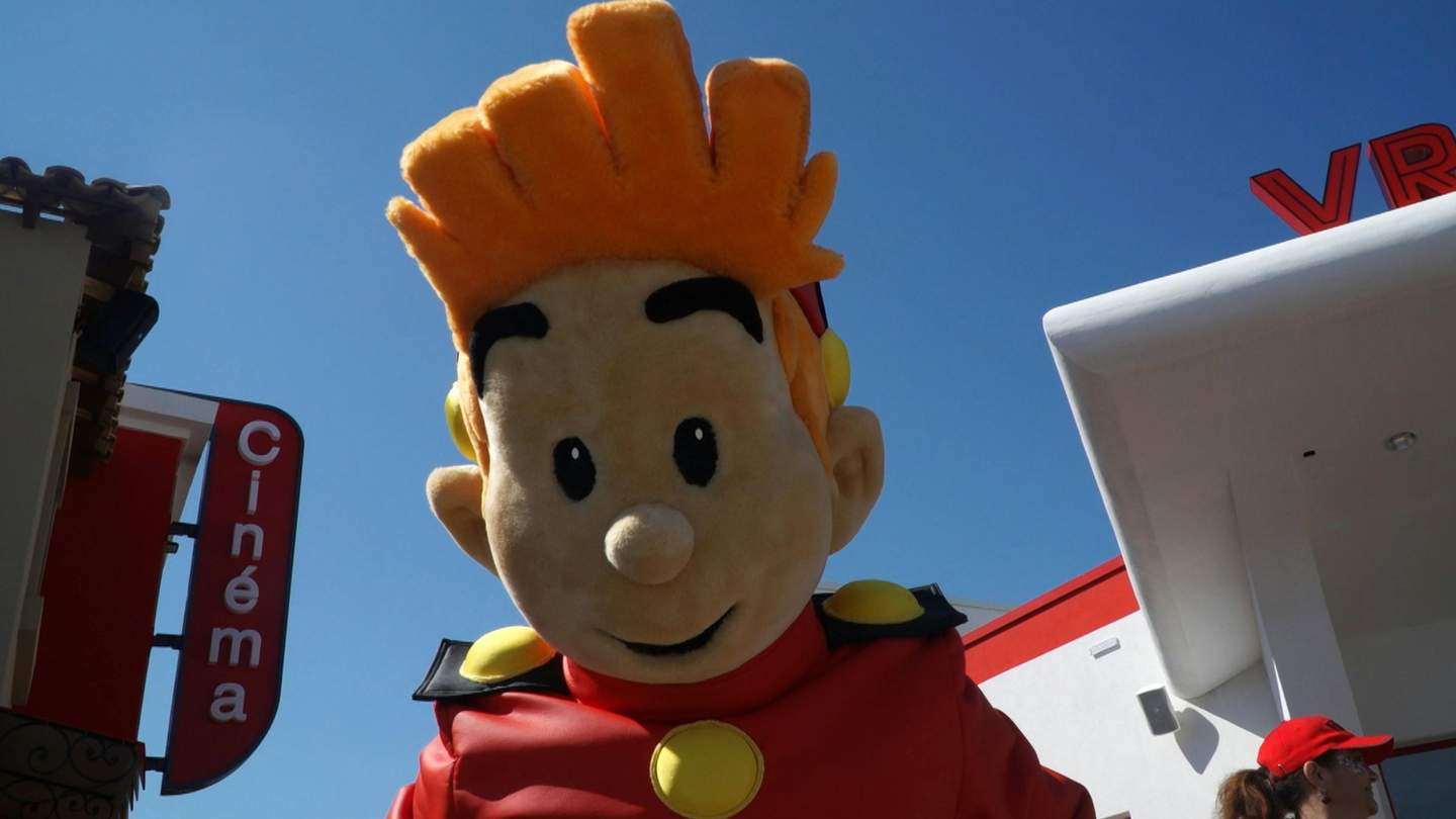 Parc Spirou Provence Large mascot with orange hair in a red outfit stands outside, next to a building with "Cinéma" sign and another white structure.