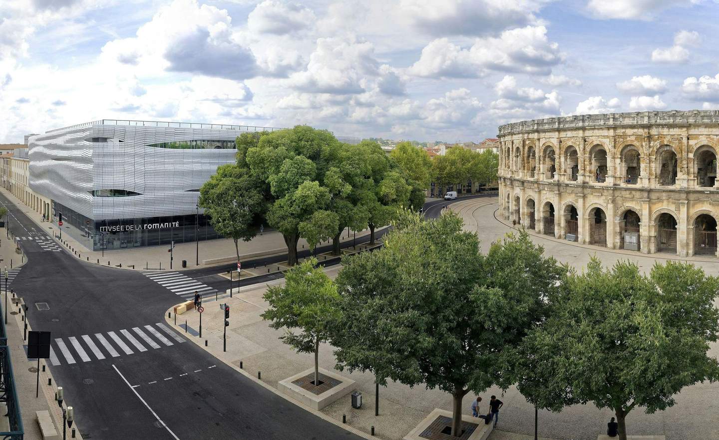 Musée de la Romanité Modern building labeled "Musée de la Romanité" next to a historic Roman amphitheater, with trees and a quiet street in the foreground.