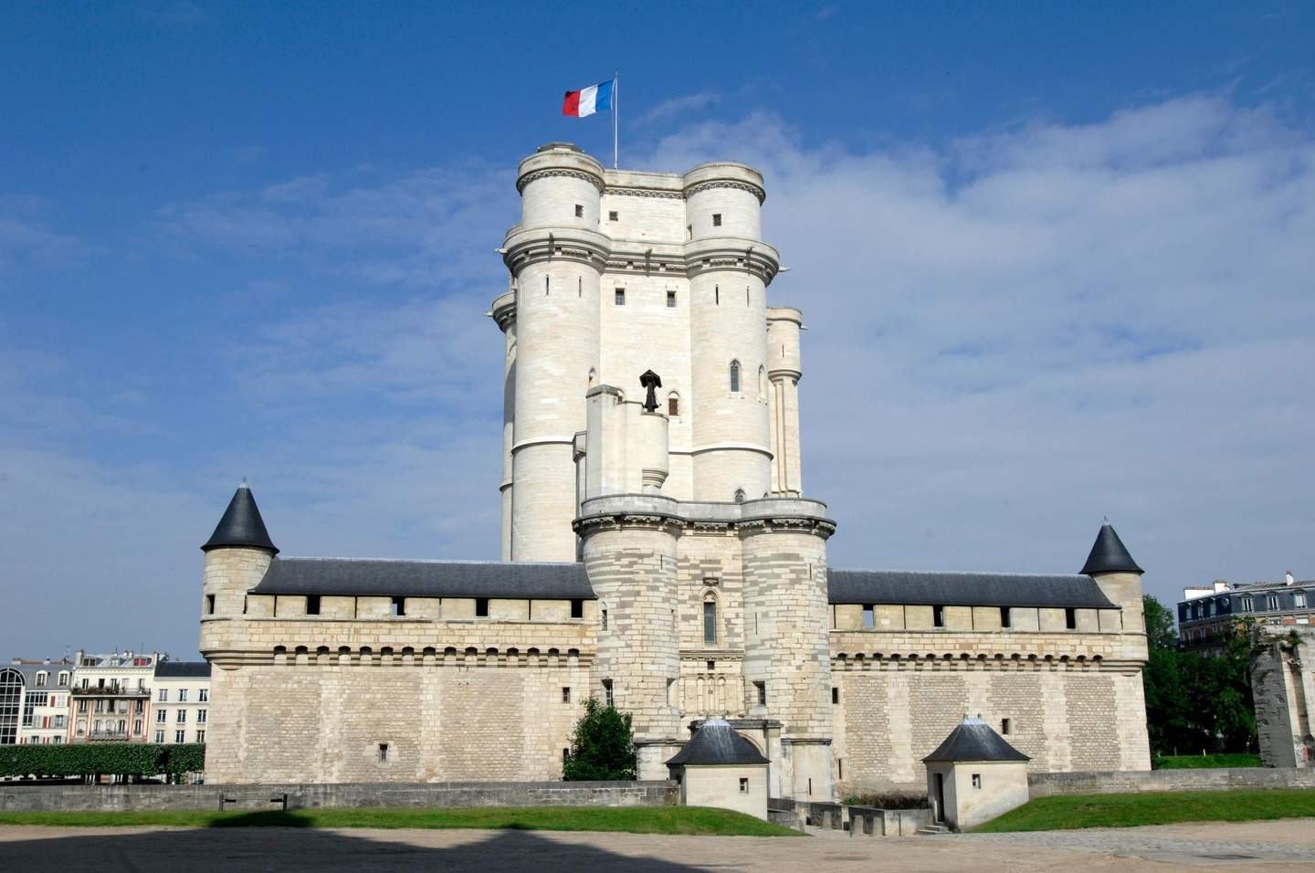 Château de Vincennes A tall, historical stone castle with turrets and a French flag on top, set under a blue sky with scattered clouds.