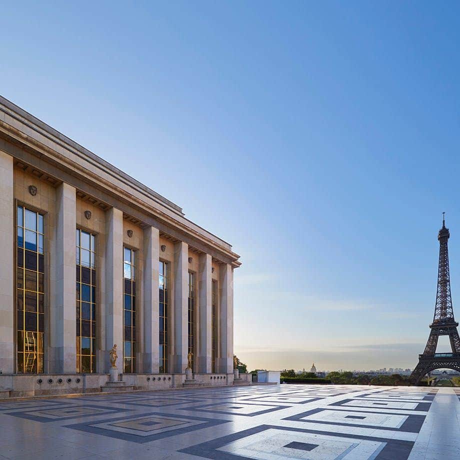 Cité de l'architecture et du patrimoine A building with columns on the left, patterned pavement, and the Eiffel Tower in the background against a clear blue sky.