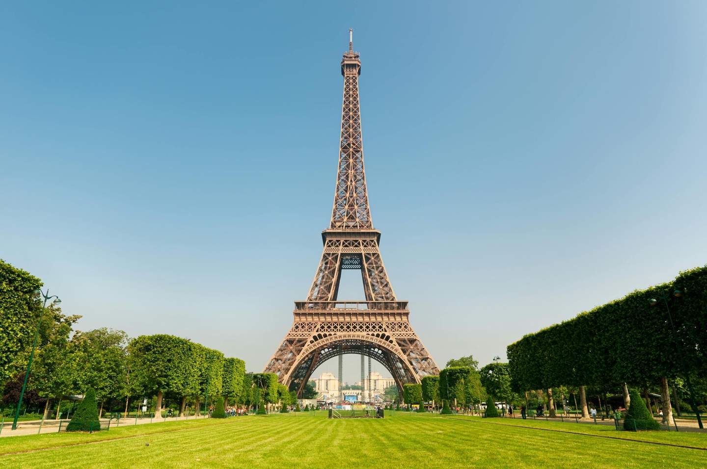 Eiffel Tower The Eiffel Tower seen from a grassy park with trimmed trees on both sides under a clear blue sky.
