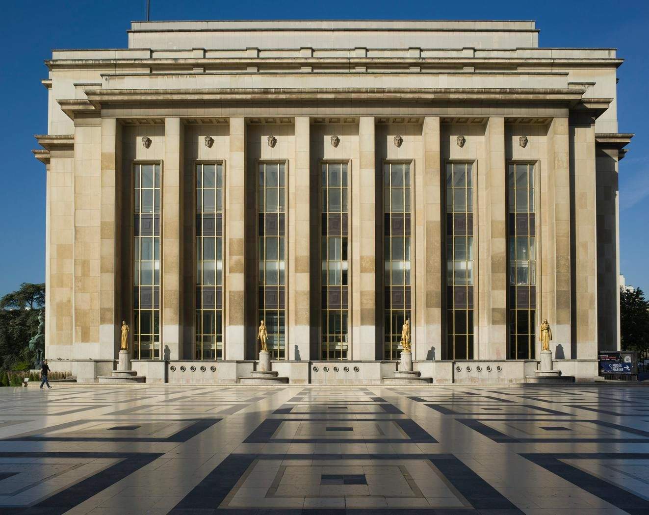 Large building with tall columns and golden statues on a geometric patterned plaza under a clear blue sky.