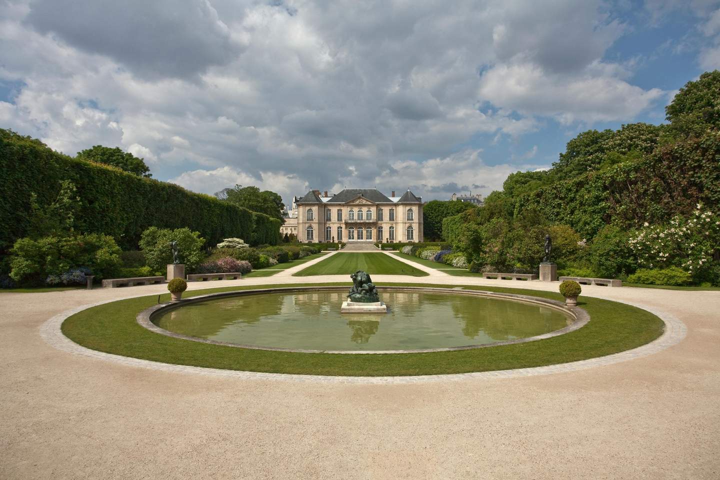 Musée Rodin Paris A grand mansion is seen through a manicured garden with a central pond featuring a statue, flanked by statues and trimmed bushes.
