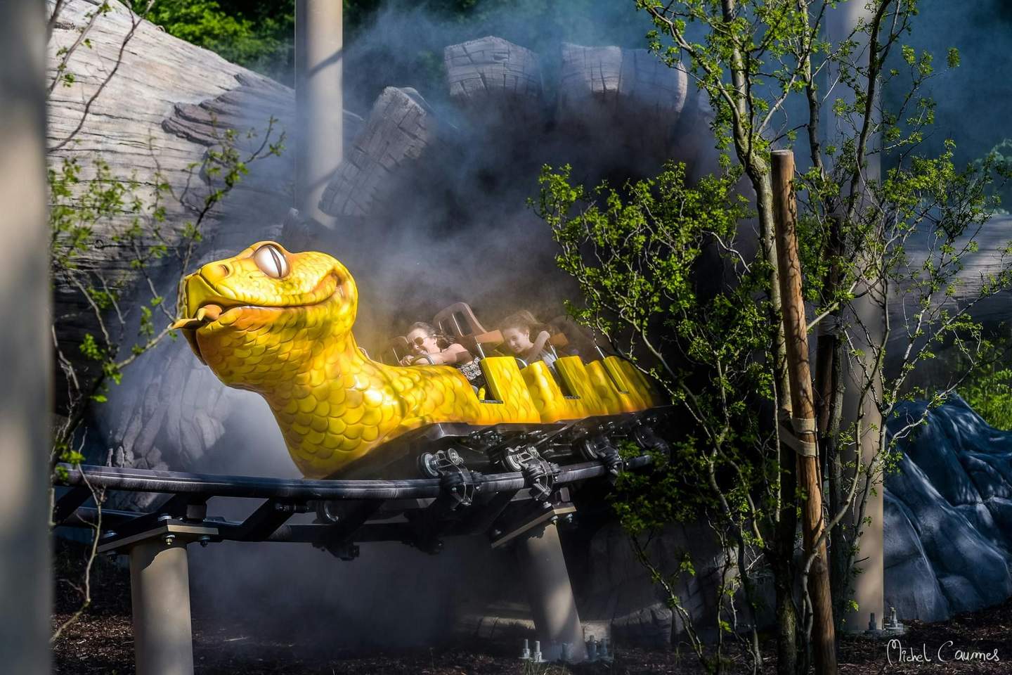 Le Parc du Petit Prince A yellow snake-themed roller coaster car with riders passes through misty, wooded surroundings.