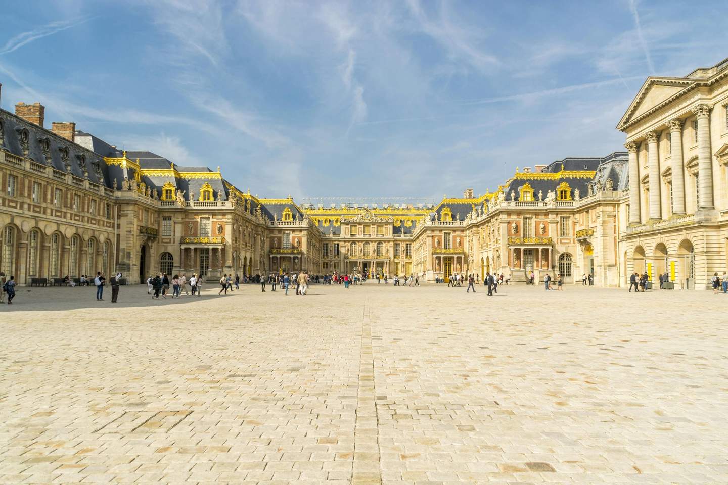 Palace of Versailles Courtyard of a grand palace with golden decorations on the rooftops. People are walking and taking photos on a sunny day.