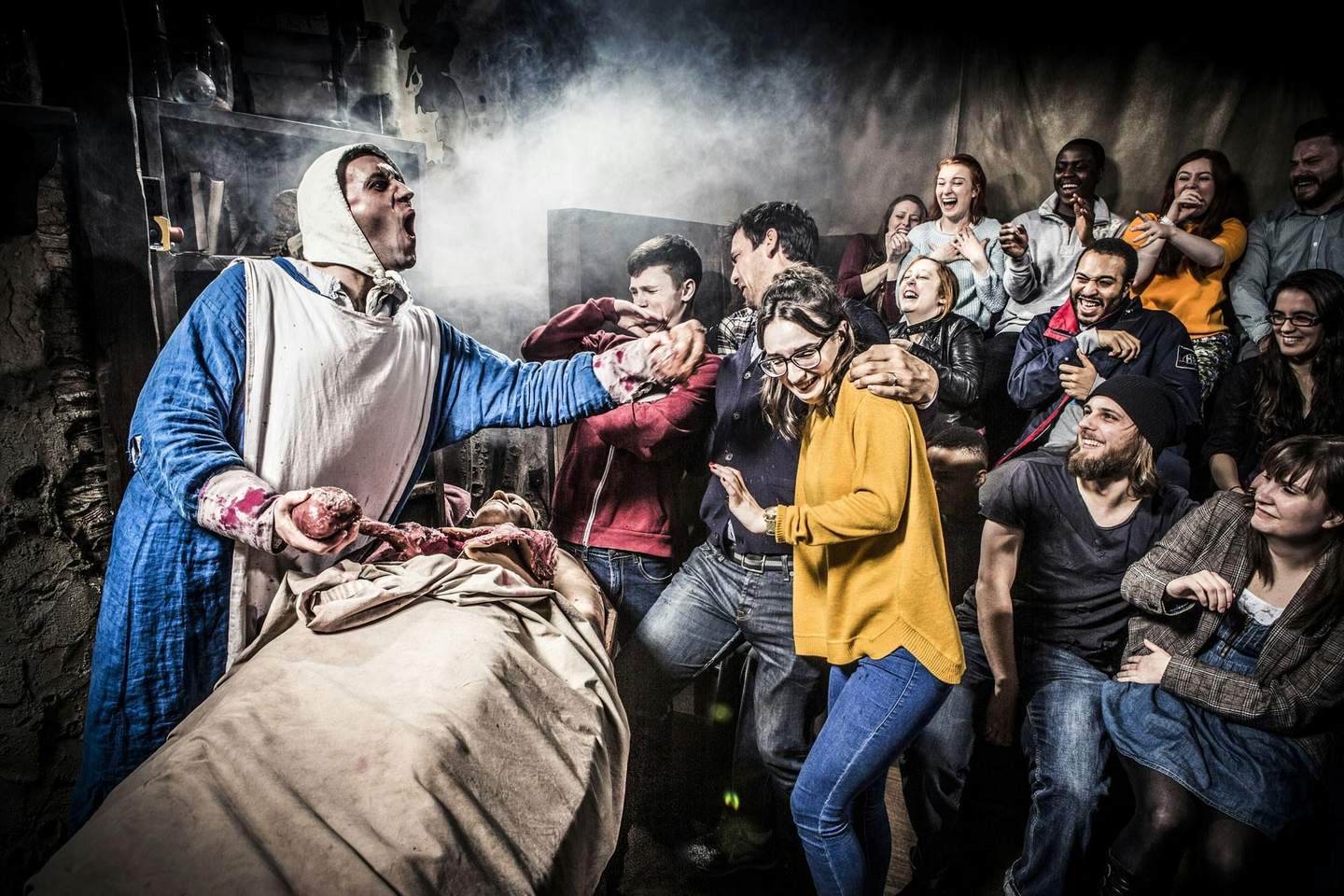 Berlin Dungeon A group of people react with amusement and shock as a costumed performer holding fake organs performs in a dark, theatrical setting with smoke.