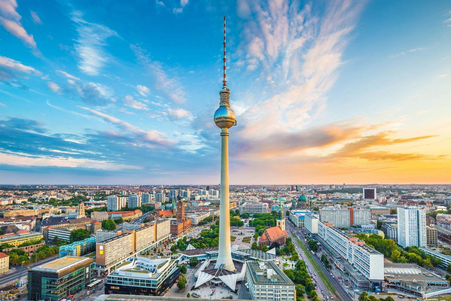 Berlin TV Tower Aerial view of a cityscape featuring a tall TV tower at the center, surrounded by buildings, streets, and greenery under a blue sky.
