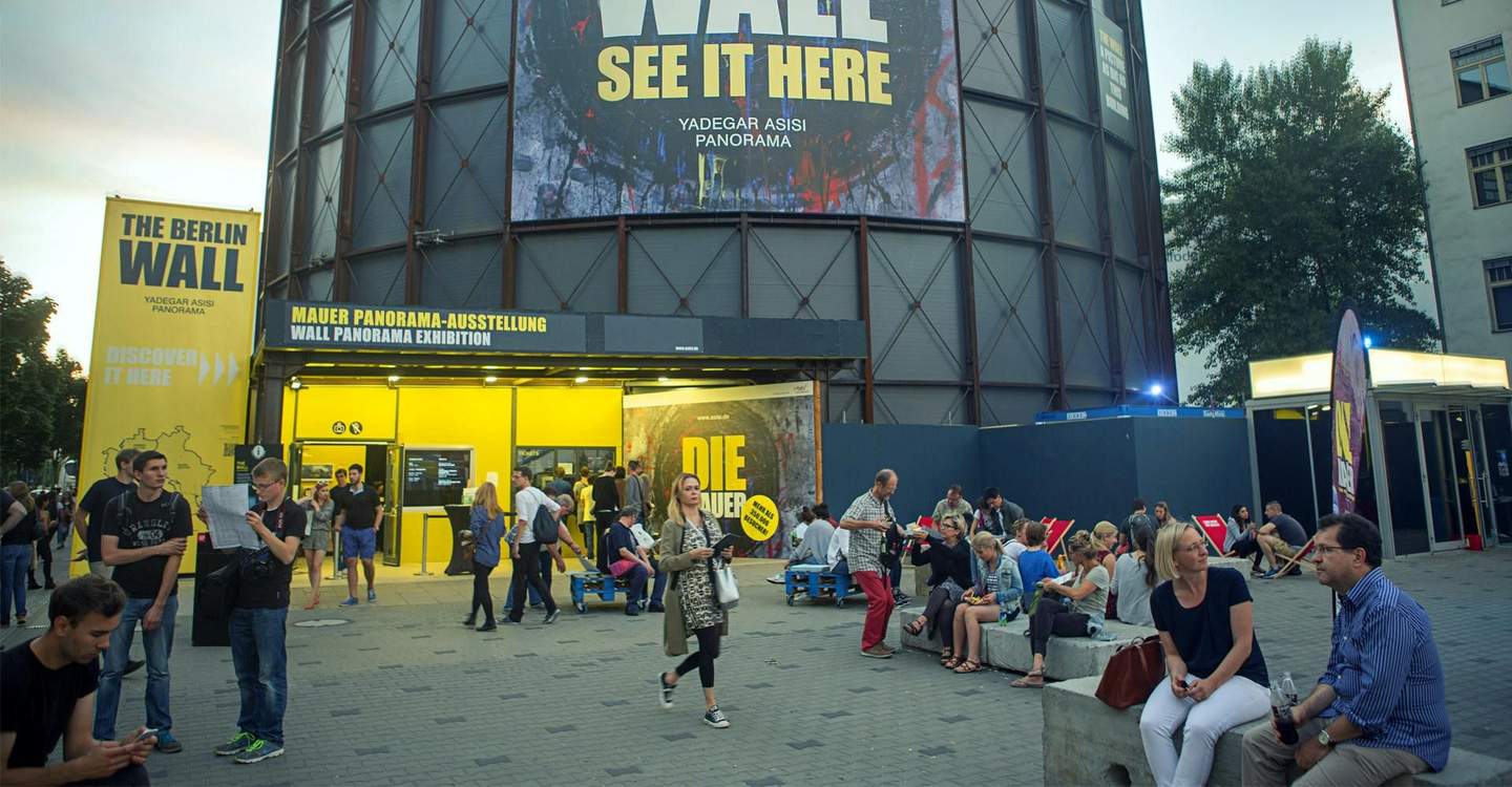 People gather outside the Berlin Wall Panorama Exhibition, with large yellow signs and posters promoting the event.
