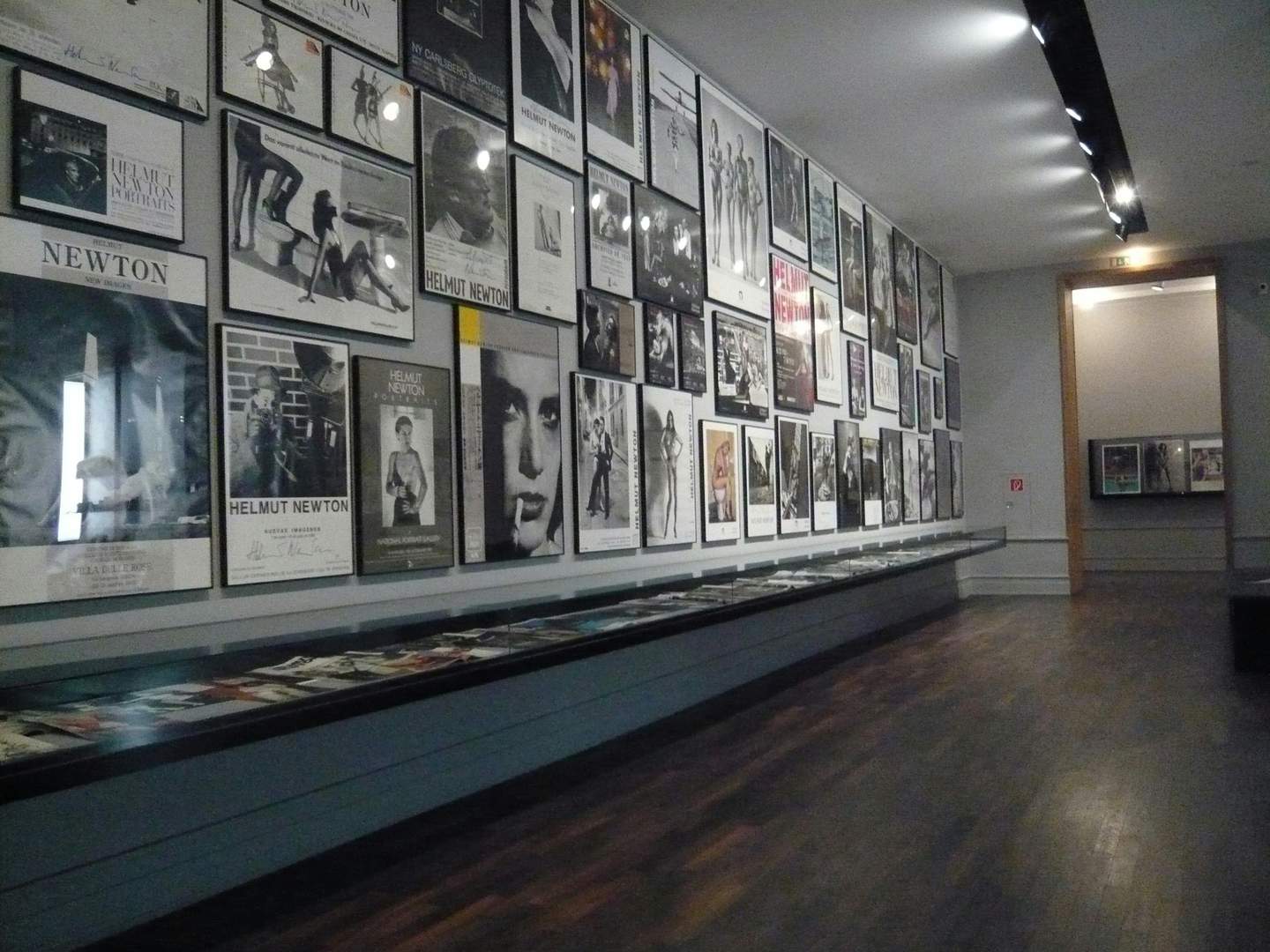 Museum of Photography / Helmut Newton Foundation A gallery wall covered with framed photographs and posters, with a glass display case below holding various printed materials.