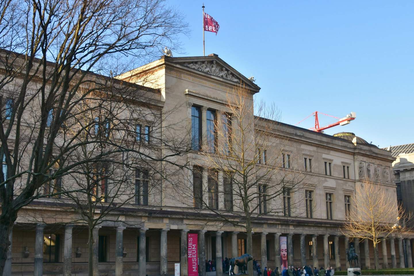 Neues Museum Large, neoclassical building with columns, a "Neues Museum" banner, and people gathered outside. Leafless trees and a red crane are visible.