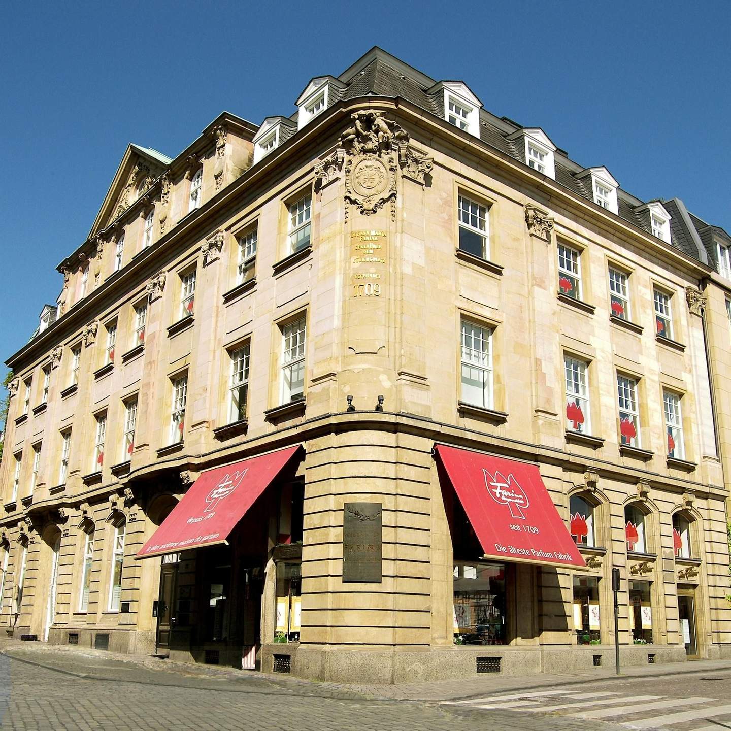 Farina Fragrance Museum Corner view of a historic beige building with red awnings and multiple windows, located on a cobblestone street under a clear blue sky.