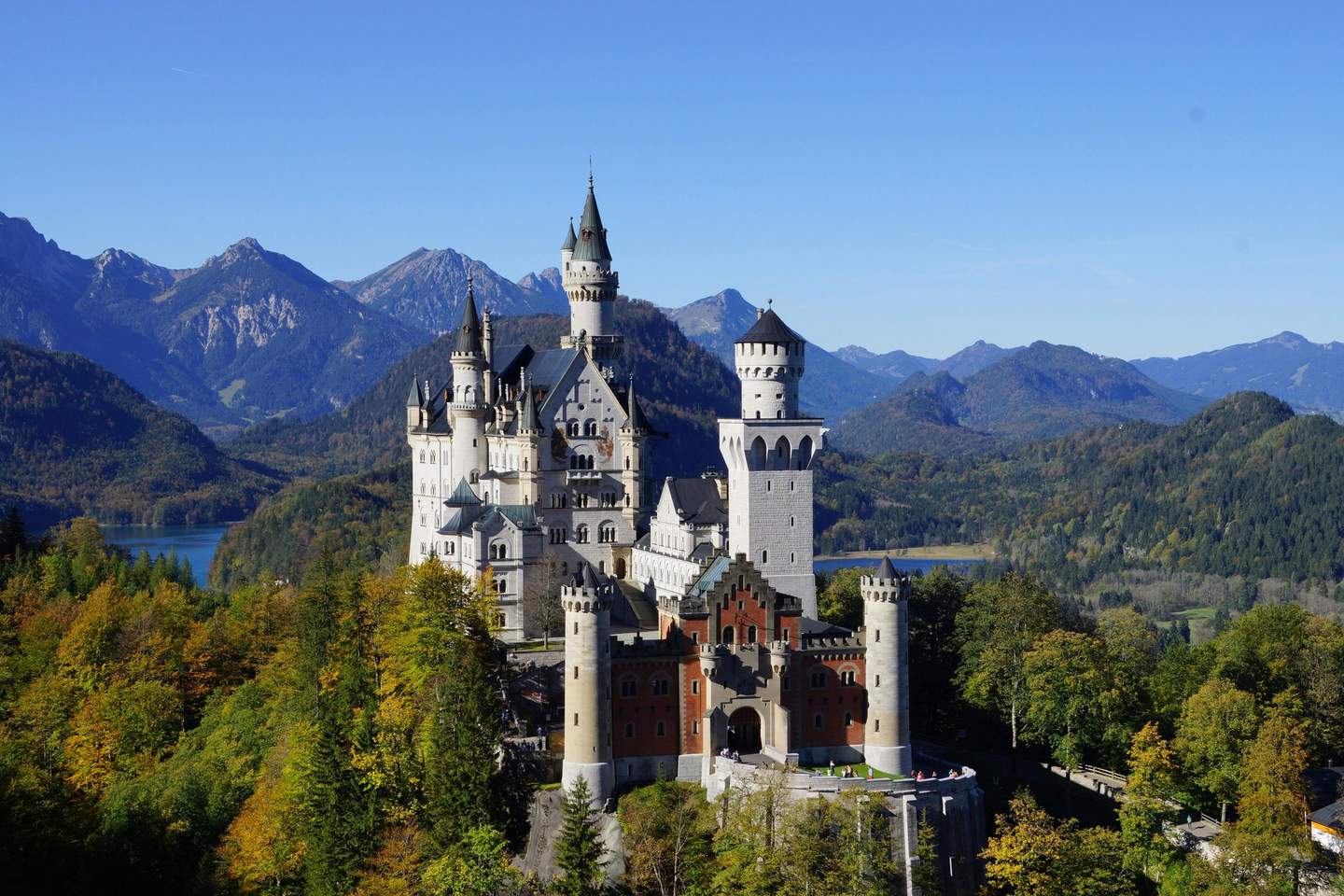 A castle with multiple towers on a forested hill, surrounded by mountains under a clear blue sky, with a lake visible in the distance.