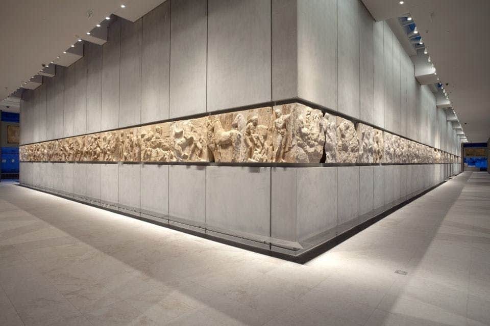 A museum exhibit features an extensive sculptural frieze, displayed along a well-lit, minimalist hallway with grey walls and ceiling lights.