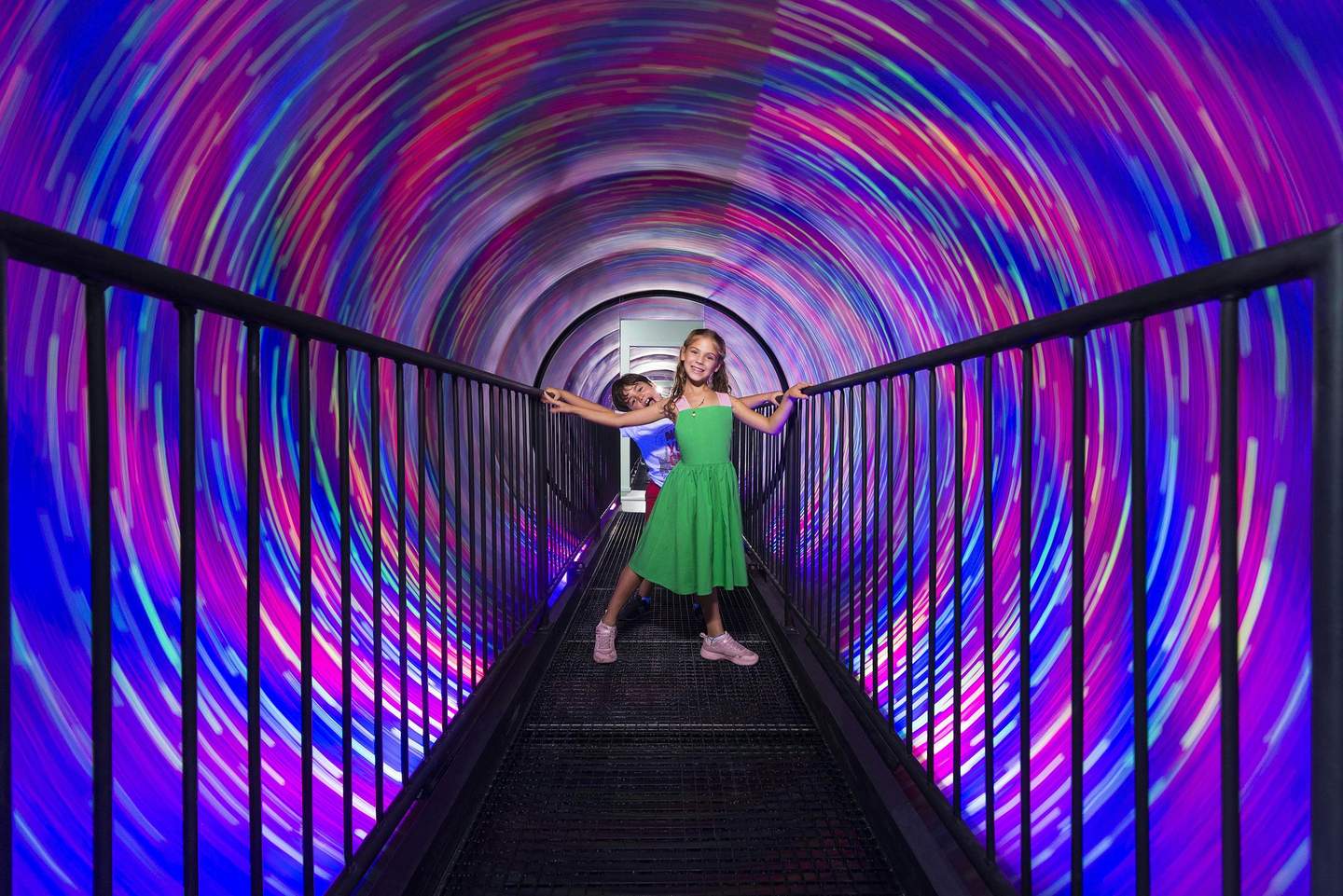 Two children stand on a walkway inside a tunnel with swirling, colorful lights. They hold onto the railings, smiling.