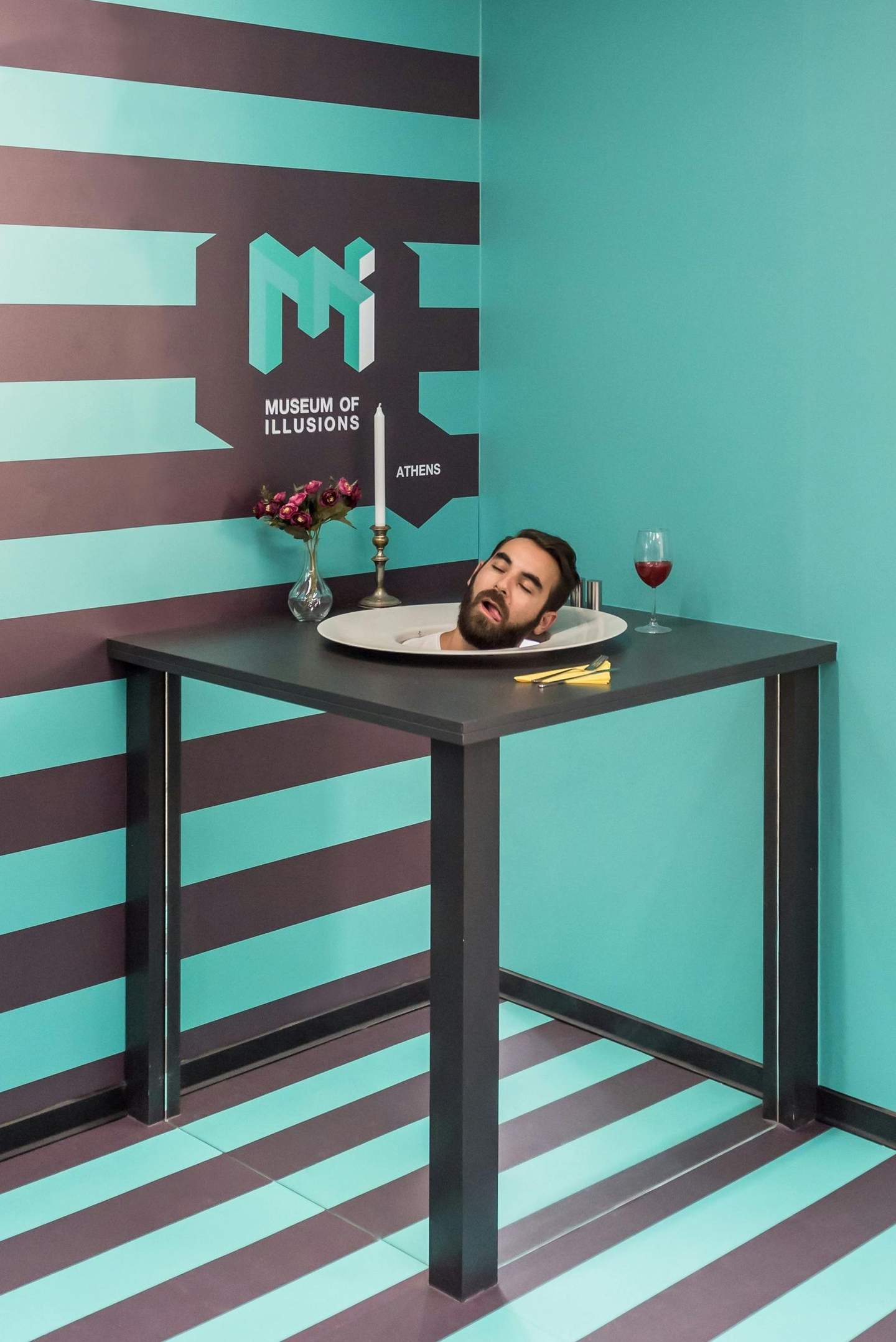 Museum of Illusions Thessaloniki A table with a man's head on a plate, a vase, a candlestick, and a wine glass, in front of a striped Museum of Illusions wall.