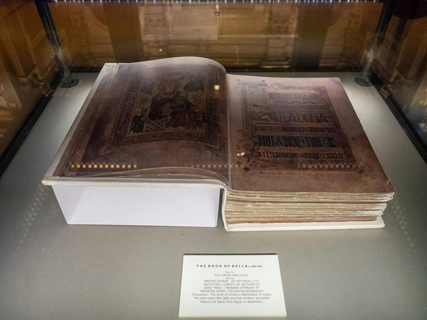 An illuminated manuscript, "The Book of Kells," displayed in a glass case with a description plaque below.