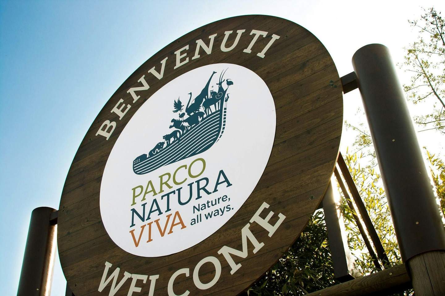 Parco Natura Viva A round wooden sign reads "BENVENUTI" and "WELCOME," featuring a logo with animals and the text "PARCO NATURA VIVA, Nature, all ways."