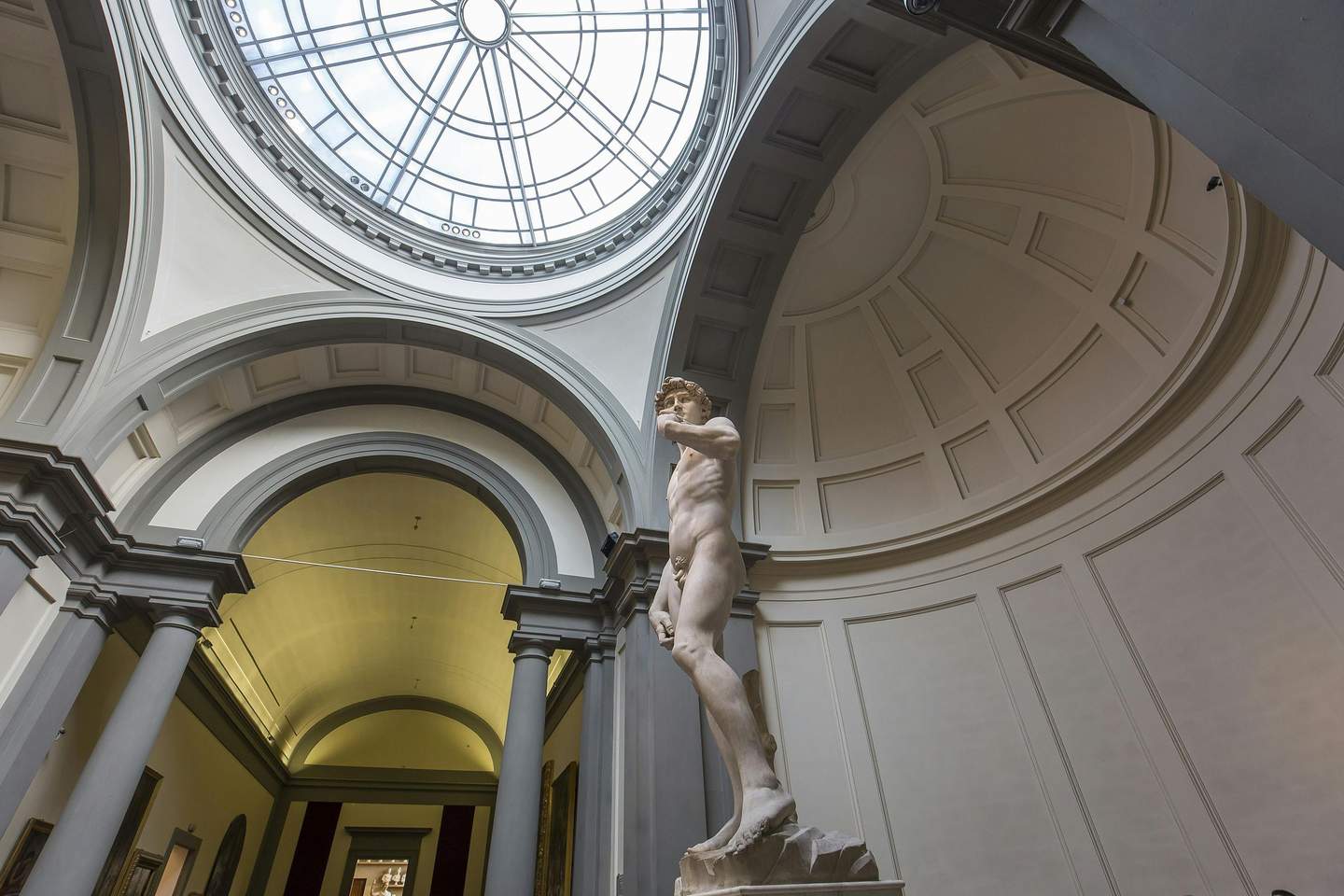 A large marble statue of a nude man stands under a domed and arched ceiling with a circular skylight.