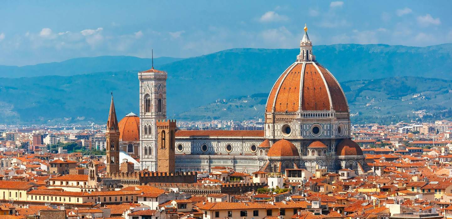 Panoramic view of Florence, featuring the Cathedral of Santa Maria del Fiore with its iconic red dome and bell tower, against a backdrop of mountains.