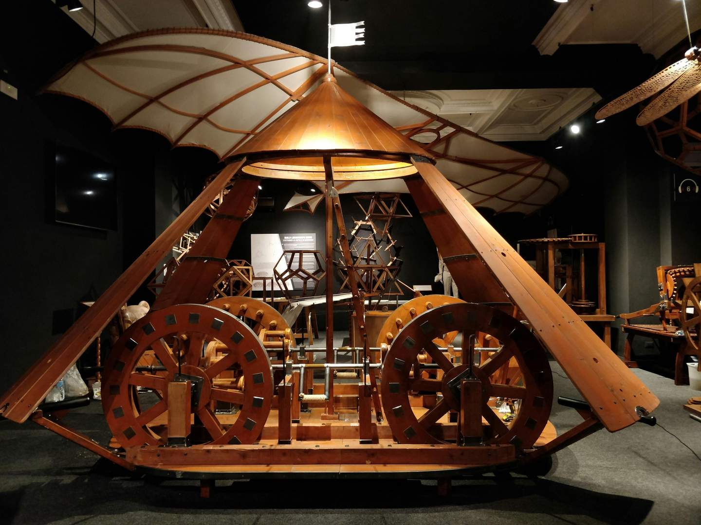 Leonardo da Vinci Interactive Museum® Large wooden machine with gears, wings, and a canopy on display in a dimly lit museum room. Other wooden models are visible in the background.