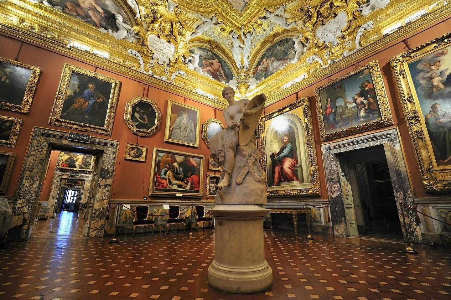 Palazzo Pitti Ornate gallery with framed paintings, a large central marble statue, gold-accented ceiling, and a checkered floor.