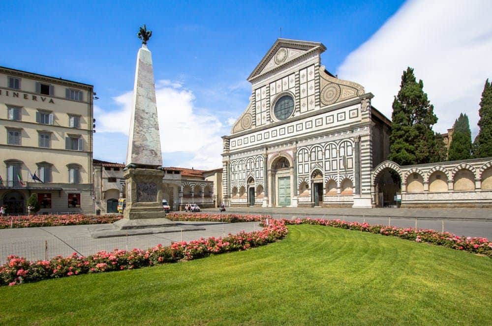 Santa Maria Novella Historical building and obelisk in a landscaped plaza with green lawn and flower beds, under a blue sky.