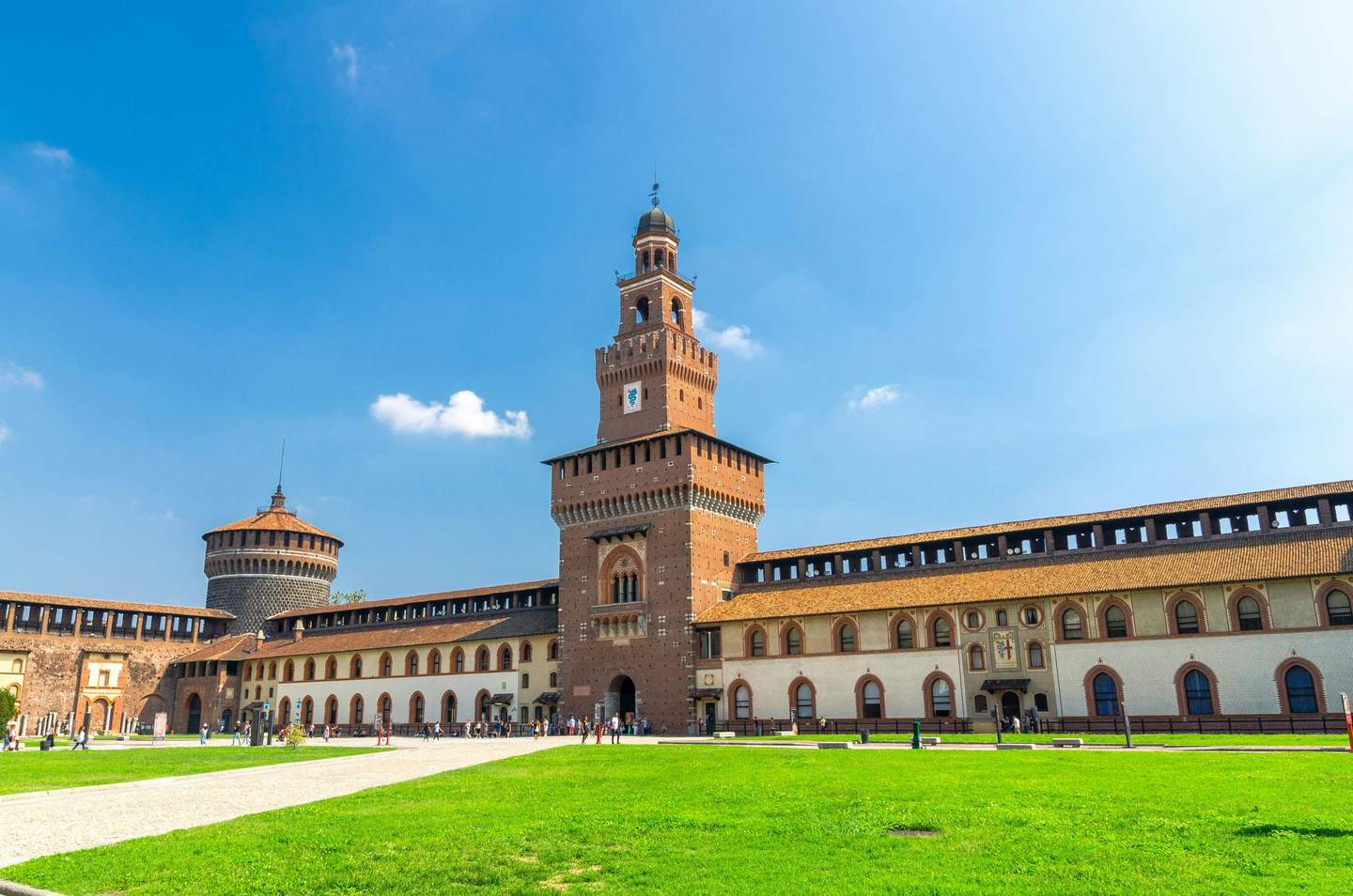 Castello Sforzesco Historic brick castle with a tall central tower, arched windows, and a green lawn, against a clear blue sky with a few clouds.