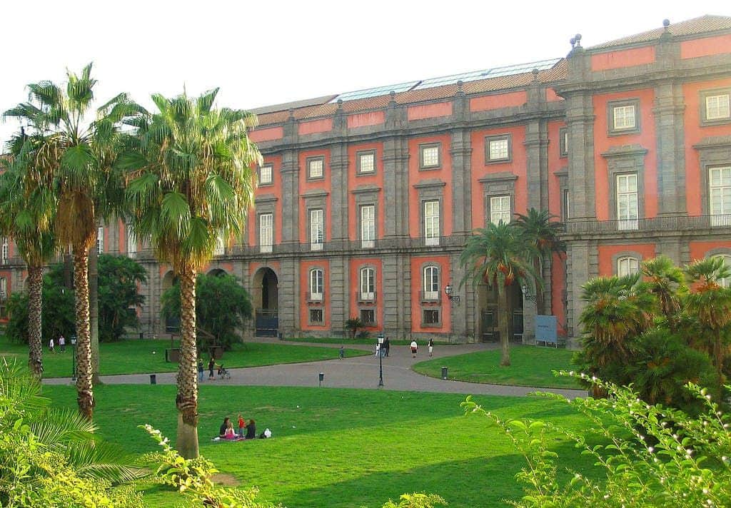 Capodimonte Museum A large red and gray building with a manicured lawn, palm trees, and people walking and sitting on the grass.
