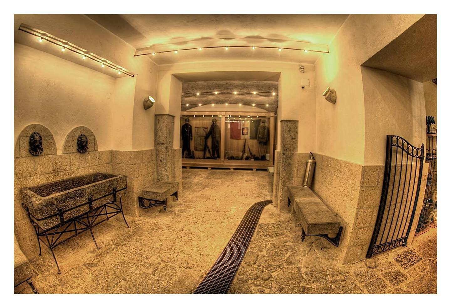 Naples Underground A well-lit, stone-walled hallway with benches, lion-shaped water fountains, and mannequins in uniforms displayed behind glass.