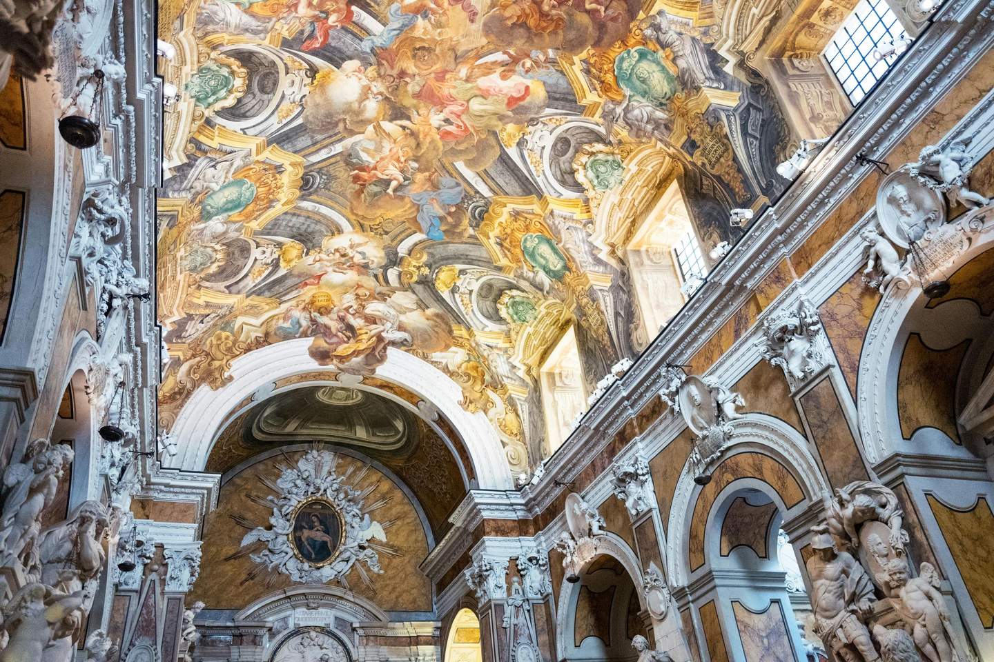 Ornate interior of a church with a detailed frescoed ceiling, archways, and statues against illuminated windows.