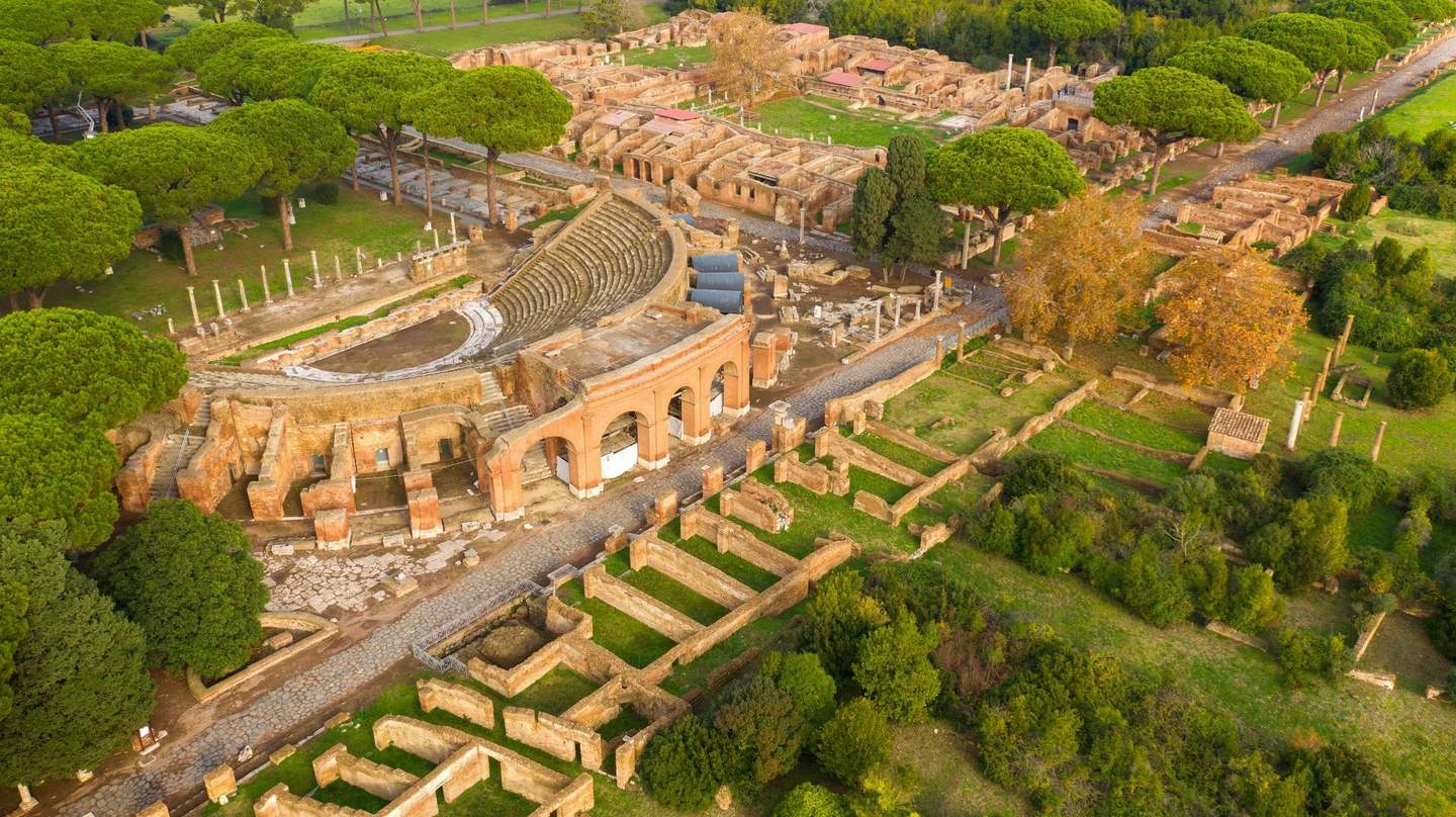 Archaeological Park of Ostia Antica Aerial view of ancient ruins including an amphitheater, columns, brick structures, and surrounding lush greenery.