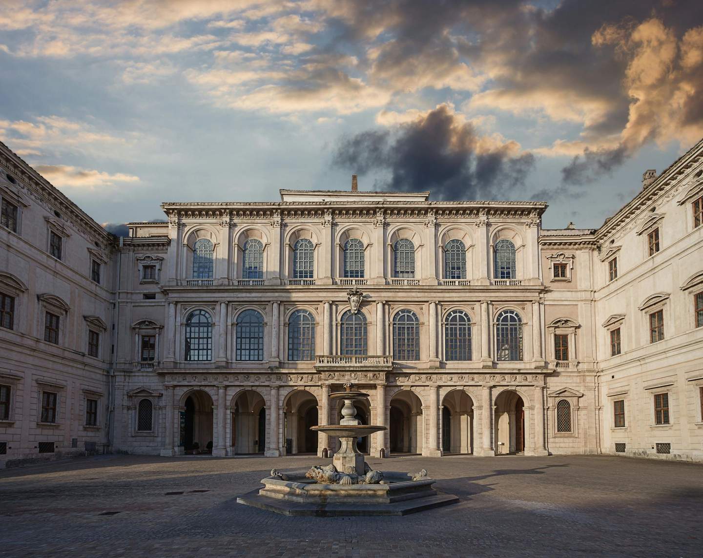 Courtyard of a historic building with arches and large windows, featuring a central fountain. Dramatic sky with clouds above.