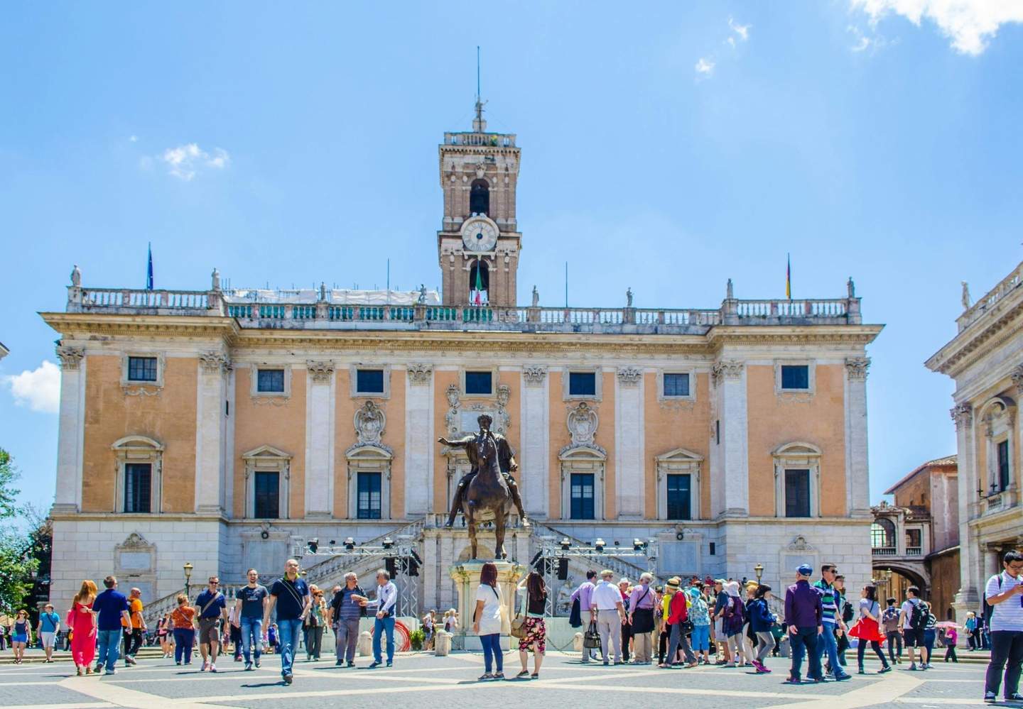 Tourists gather around a statue on a horse in front of an ornate, historic building with a clock tower under a clear sky.