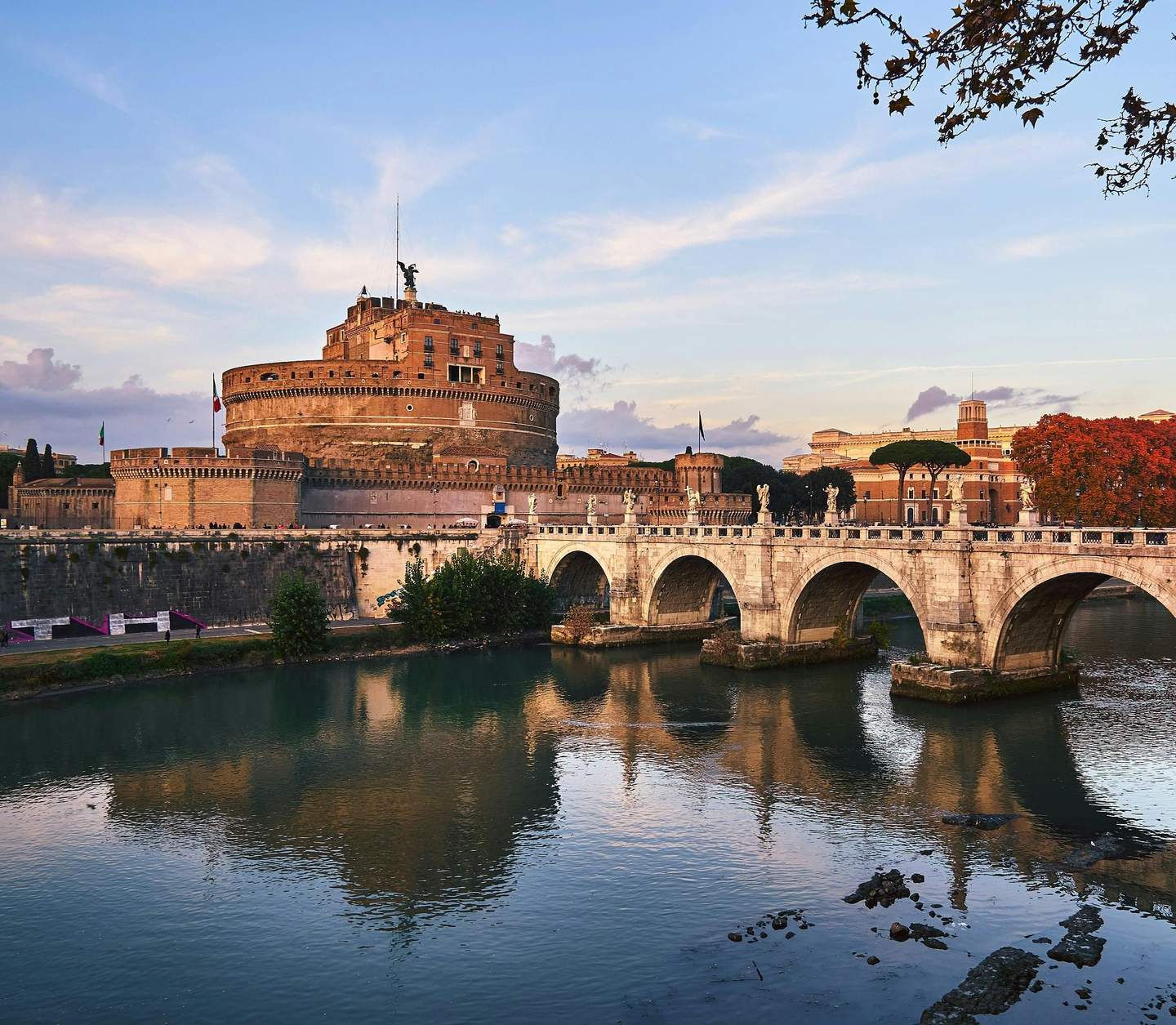 Historic stone bridge with arches spans a river, leading to a large cylindrical castle. Autumn trees and buildings in the background.