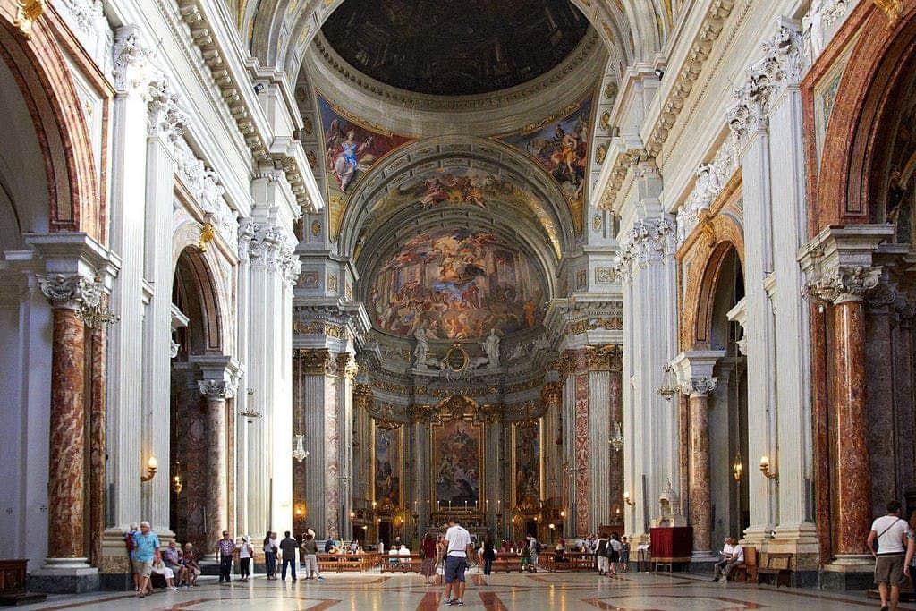 People walking inside a grand cathedral with intricate columns, arches, and detailed ceiling paintings.