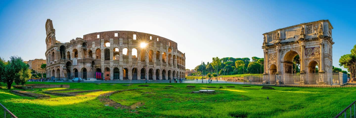 Colosseum Ancient Colosseum and Arch of Constantine in Rome, with green grass and scattered visitors under a clear blue sky.
