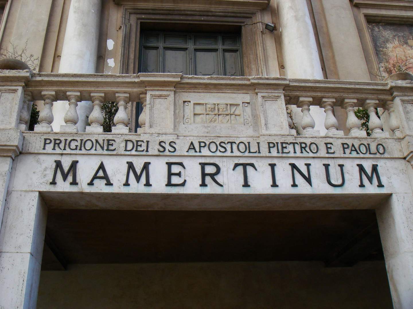 A stone entrance with a balustrade above it, featuring the text "PRIGIONE DEI SS APOSTOLI PIETRO E PAOLO MAMERTINUM."