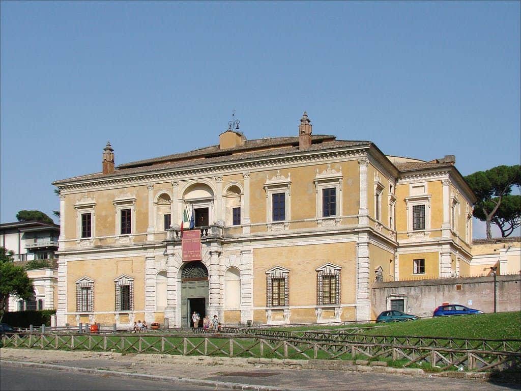 Large yellow historic building with arched entrance, stone detailing, and barred windows. Few people and parked cars outside. Clear sky.