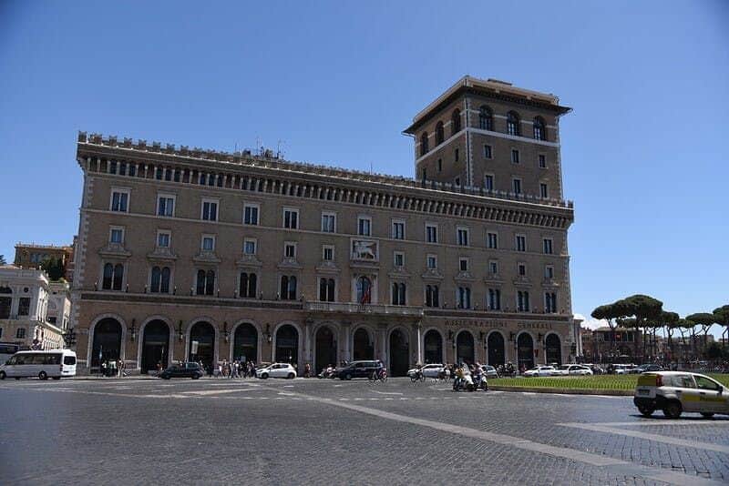 Palazzo Venezia A large historic building with arches, vehicles, and people in front, under a clear blue sky. Trees and smaller buildings in the background.