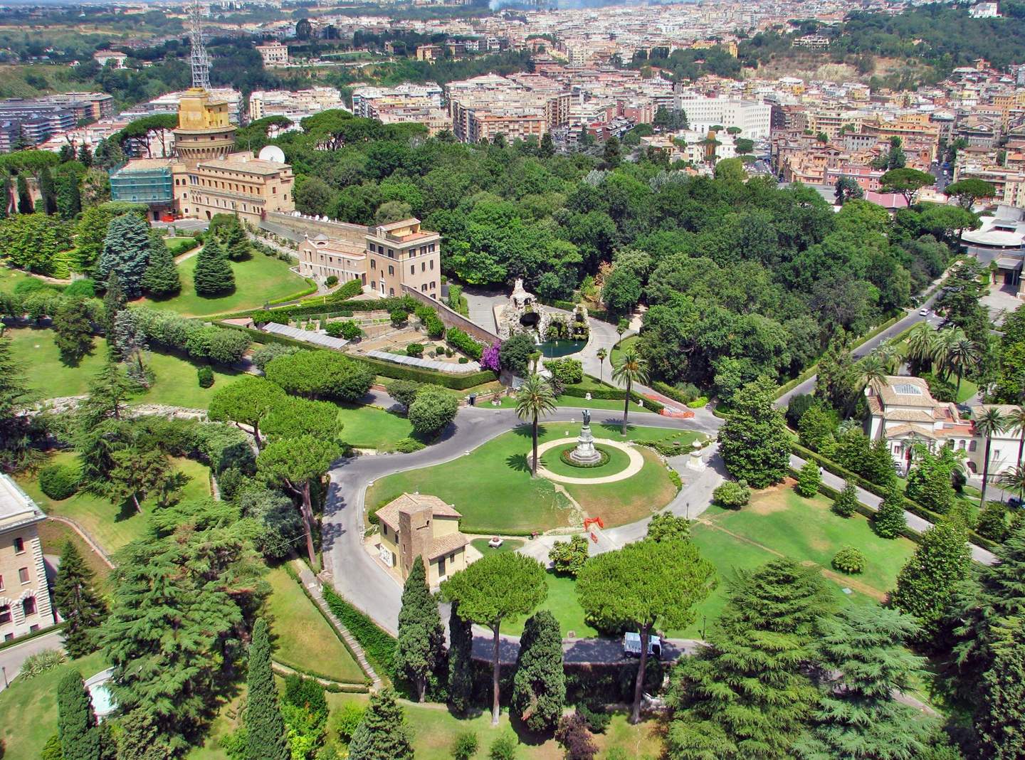 Vatican Gardens Aerial view of lush gardens with trees, winding paths, buildings, a fountain, and cityscape in the distance.