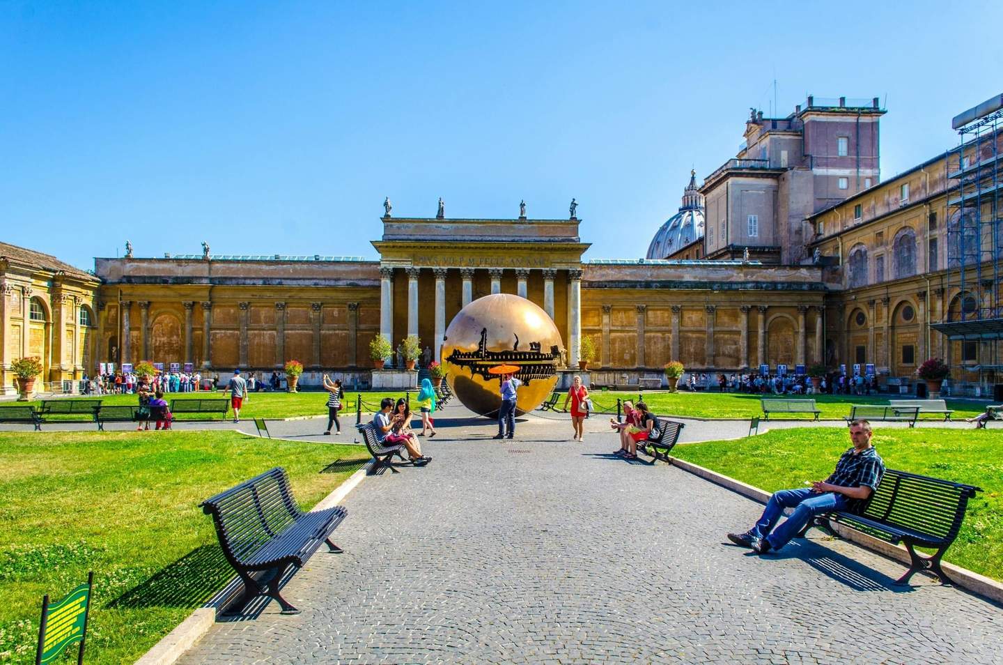 Vatican Museums Tourists relax on benches in a courtyard with a large bronze sculpture and historic building under a clear blue sky.