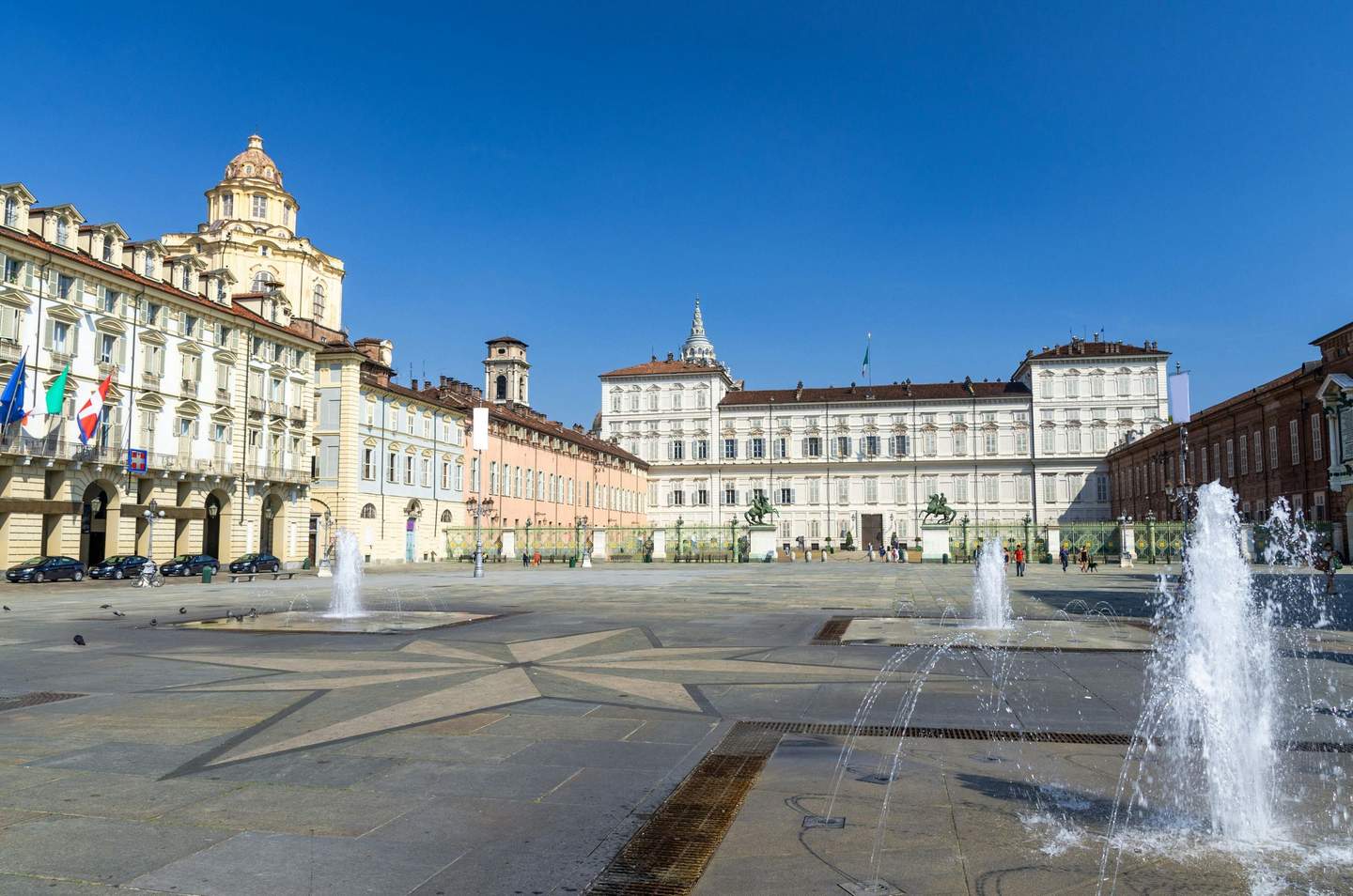Musei Reali Torino A large, open square with several fountains, historic buildings with flags, and few people under a clear blue sky.