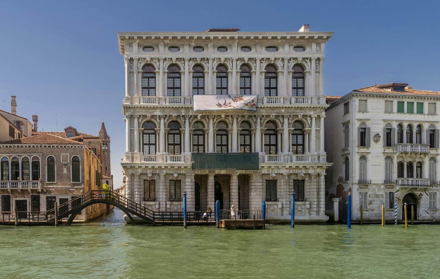 Ca' Rezzonico Museum A large, ornate building by a canal with a banner, surrounded by smaller buildings and a wooden footbridge over the water.