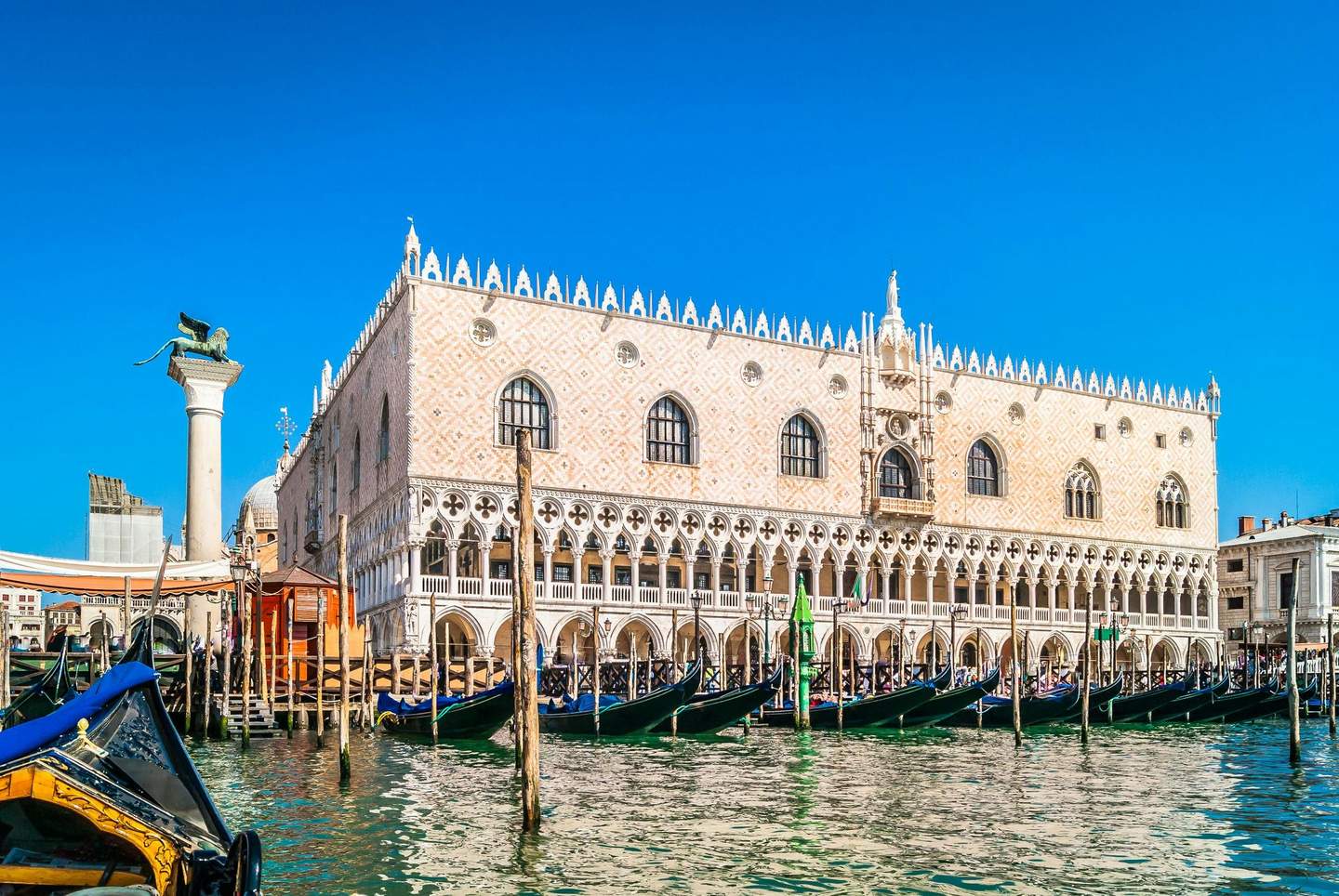 Doge's Palace Gondolas docked on a canal in front of a historic building with arched windows and a statue-topped column under a clear blue sky.