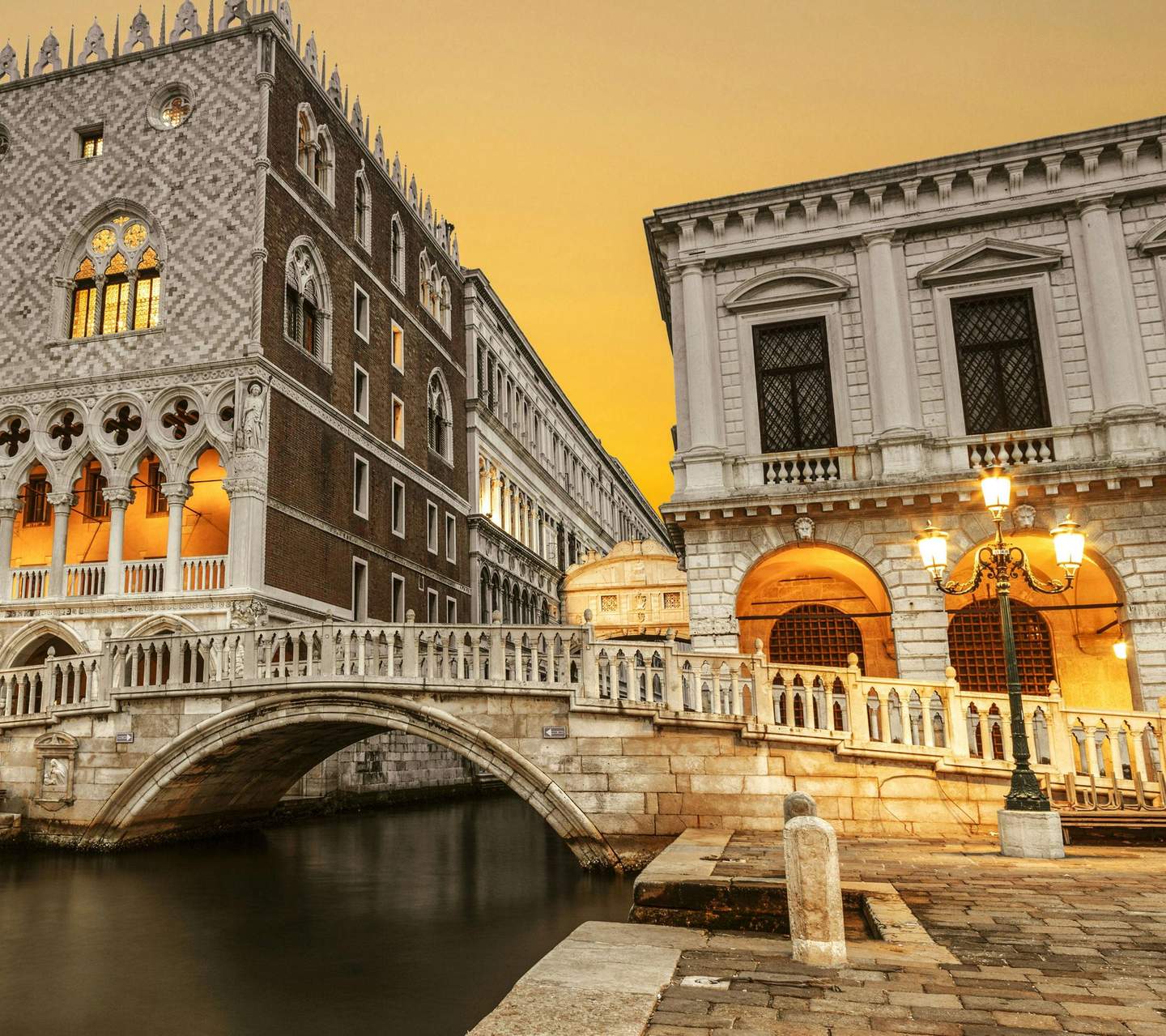 Ornate bridge and historical buildings along a canal at sunset, with lit street lamps and warm yellow windows.