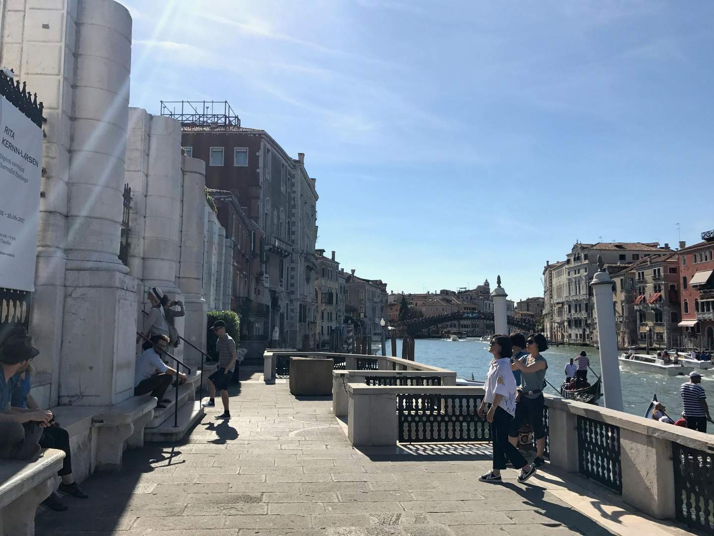 Peggy Guggenheim Collection People walk and rest by a canal in Venice with boats and historic buildings in the background under a clear, blue sky.