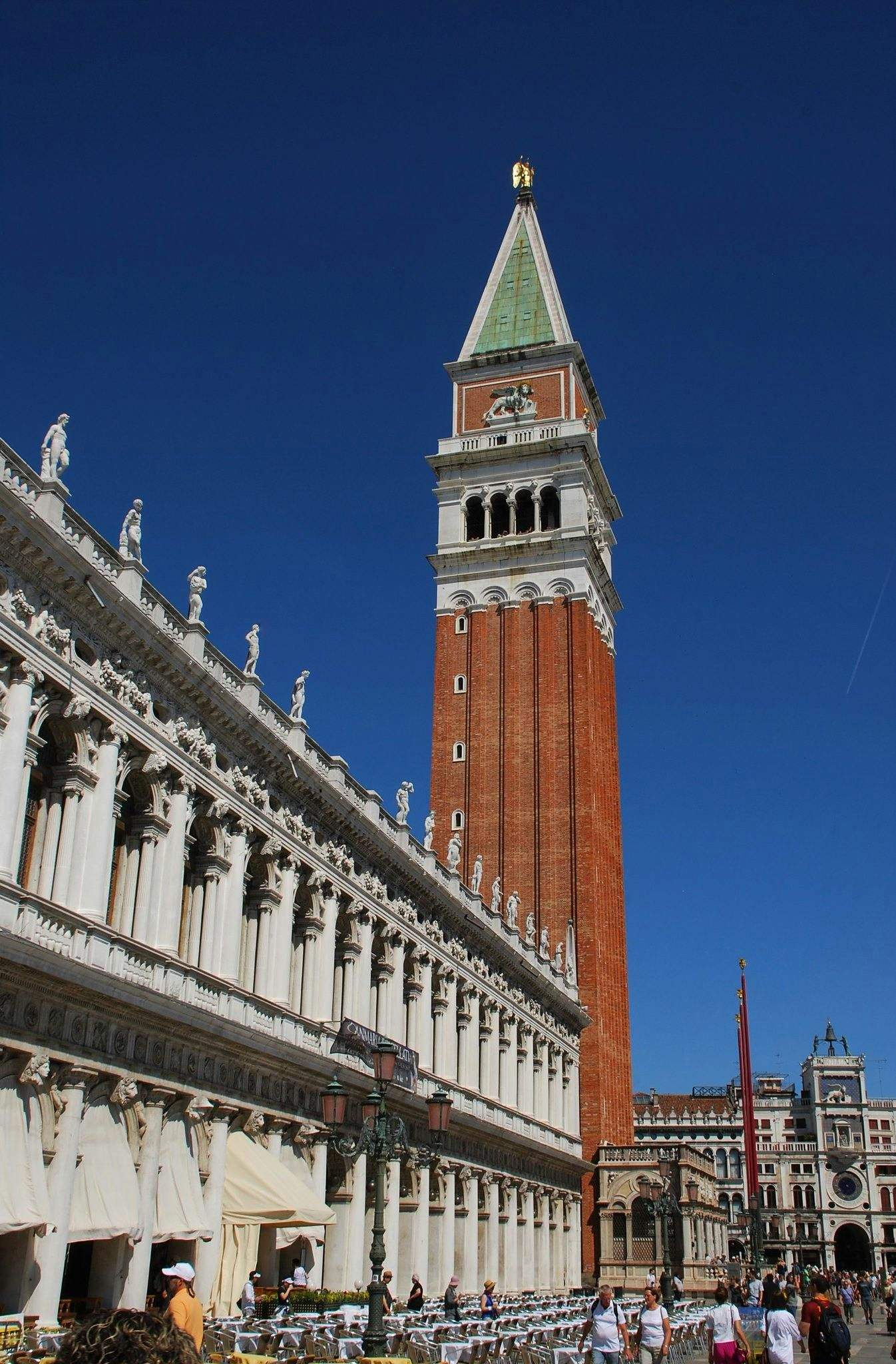 Tall, red-brick bell tower with a green spire beside a white building adorned with statues in a busy square with people and cafés.