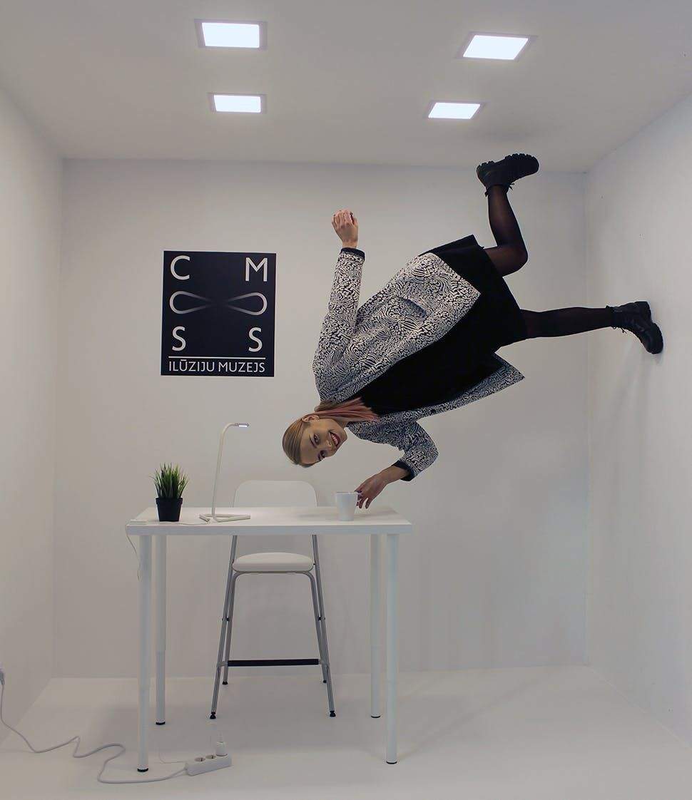 A person appears to float sideways in a minimalist room with a white table, chair, lamp, small plant, and a sign reading "ILŪZIJU MUZEJS."