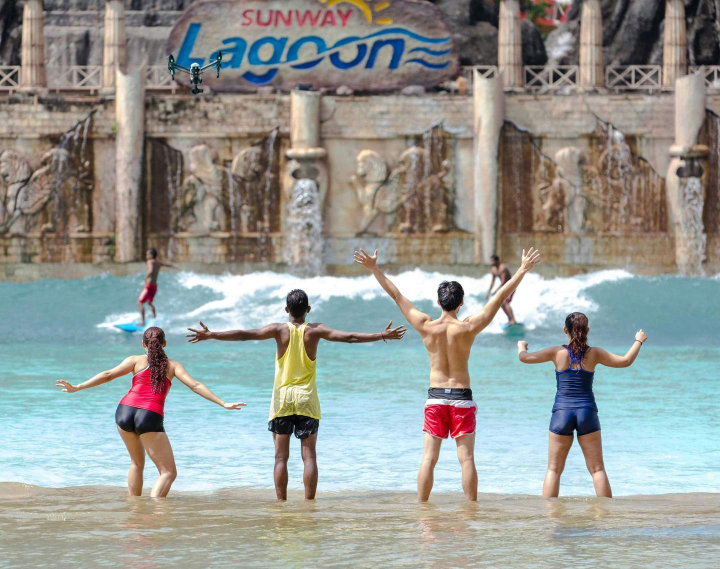 Sunway Lagoon Four people in swimwear stand in shallow water with arms raised, facing a wave pool at Sunway Lagoon. A surfer rides a wave.