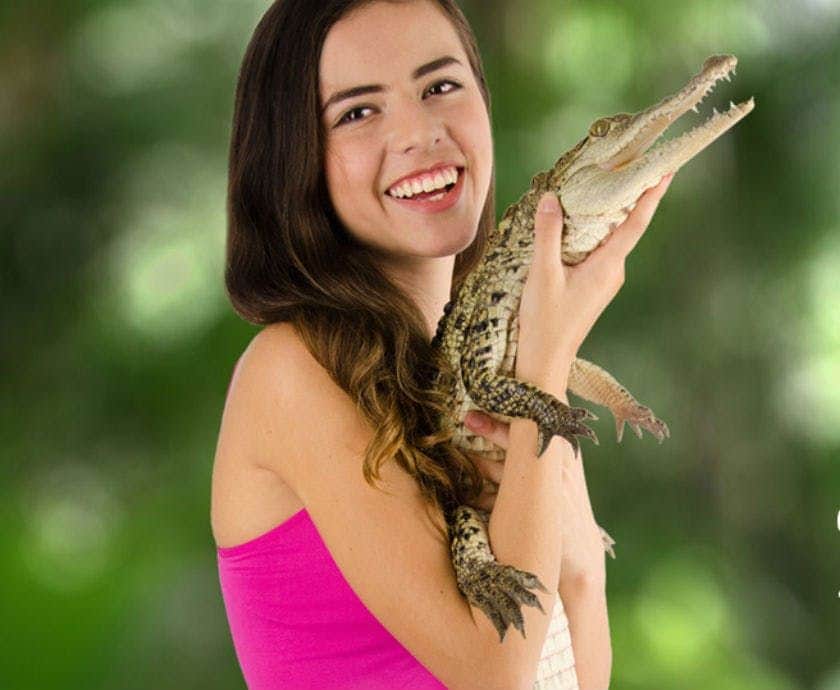 Croco Cun Zoo Smiling person in a pink top holds a small crocodile with a blurred green background.